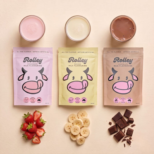 Three stand-up pouches of Rolley milk flavouring in pastel pink, yellow, and beige, arranged on a cream background with corresponding prepared drinks above and fruit/cheese toppings below. The pouches show the Rolley brand and cow illustrations.